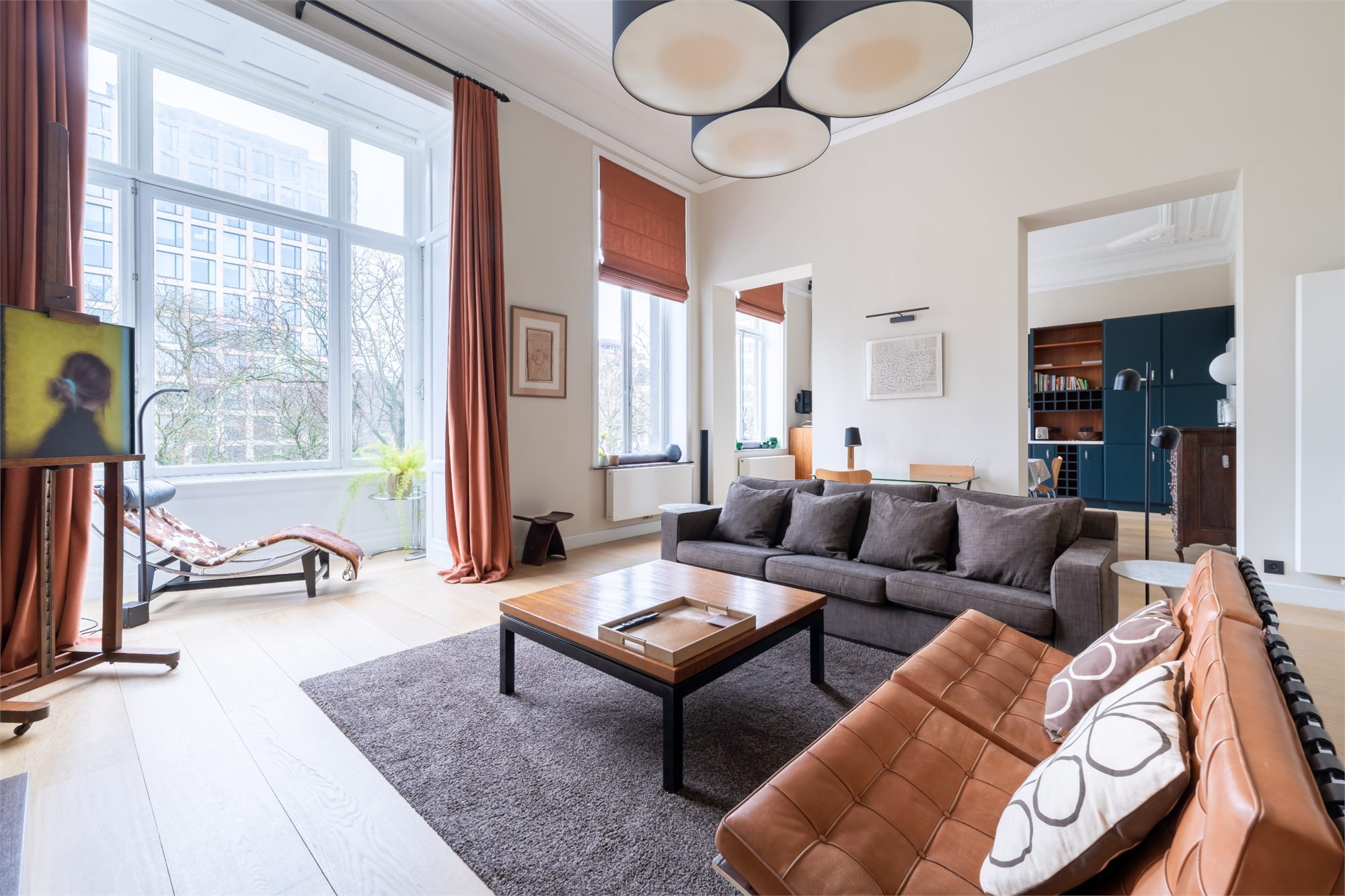 Grand salon haut sous plafond avec hautes fenêtres, canapé gris, canapé en cuir cognac et chaise longue design
