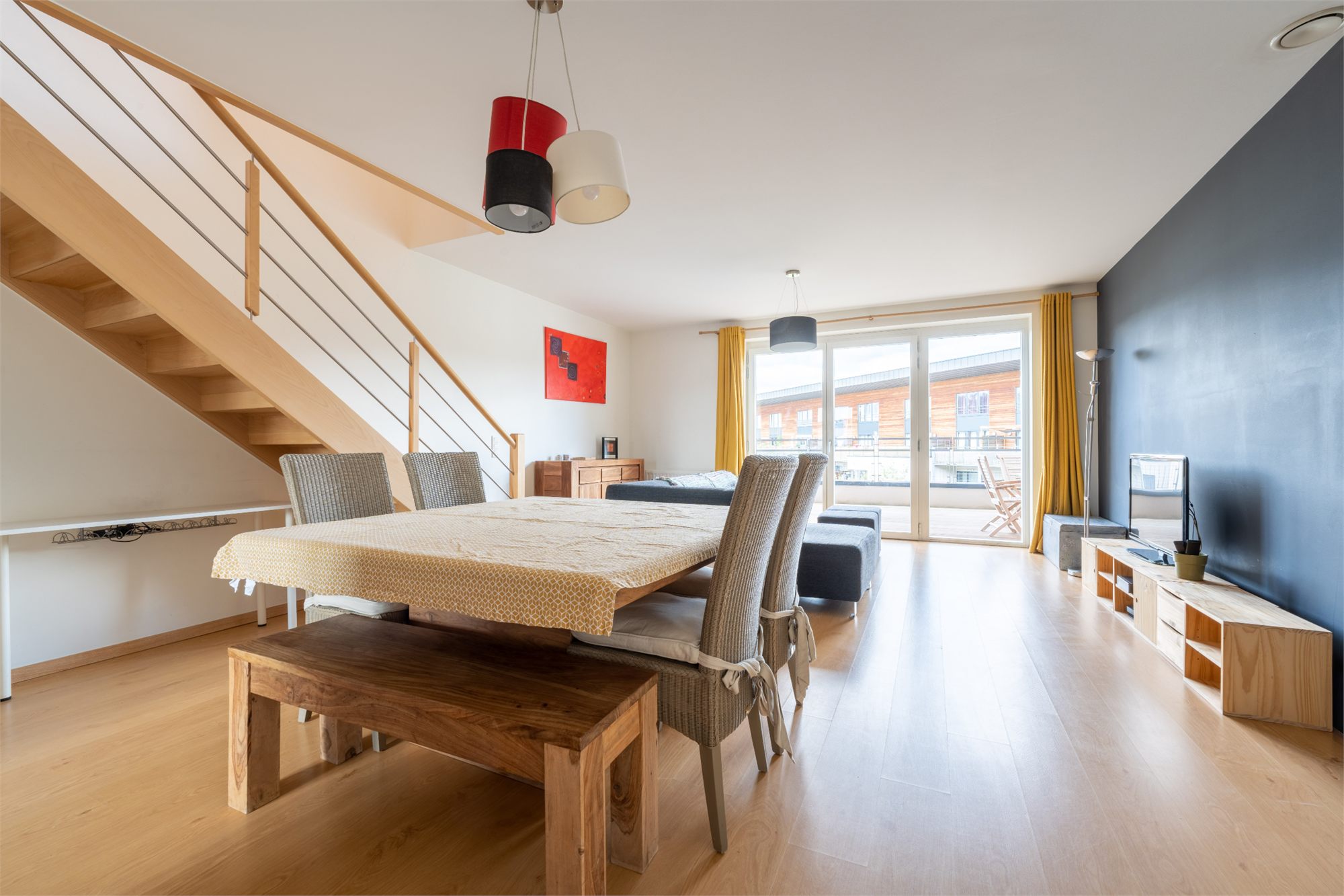 Pièce de vie en duplex avec escalier en bois, grande table et baie vitrée ouverte sur une terrasse